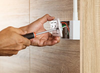 Electrician is using a screwdriver to install a power outlet in to a plastic box on a wooden wall.
