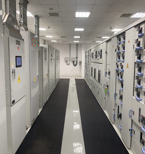 A long room with two rows of electrical panels with a black mat down the center.
